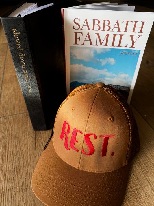 Brown cap with 'REST.' in red text, 'Sabbath Family' journal, and black book on a wooden surface.